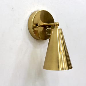 May include: A brass wall sconce with a conical shade and a circular backplate. The sconce has a pivoting arm and a polished finish. The design is minimalist and modern, suitable for interior lighting.