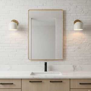May include: A modern bathroom vanity with a rectangular mirror in a gold frame. The vanity features a white countertop, a black faucet, and light wood cabinets with black hardware. Two white sconces with gold accents flank the mirror, illuminating the space.