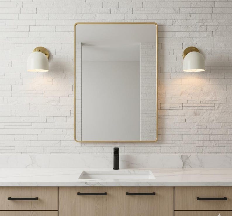 May include: A modern bathroom vanity with a rectangular mirror in a gold frame. The vanity features a white countertop, a black faucet, and light wood cabinets with black hardware. Two white sconces with gold accents flank the mirror, illuminating the space.