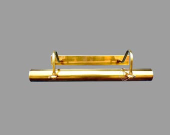 May include: A brass picture light fixture with a cylindrical body and a rectangular backplate. The fixture has two curved arms that connect the body to the backplate. The finish is a shiny gold color.