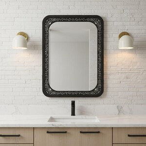 May include: A black rectangular mirror with a decorative border hangs above a bathroom vanity. Two white and gold sconces flank the mirror. The vanity has light wood cabinets with black hardware and a white countertop.
