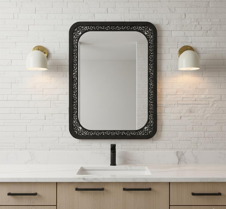 May include: A black rectangular mirror with a decorative border hangs above a bathroom vanity. Two white and gold sconces flank the mirror. The vanity has light wood cabinets with black hardware and a white countertop.