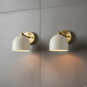 May include: Two matching wall sconces with a cream-colored, dome-shaped shade and a brass-colored circular backplate. The sconces are mounted on a gray wall, and the light from the bulbs creates a warm glow.