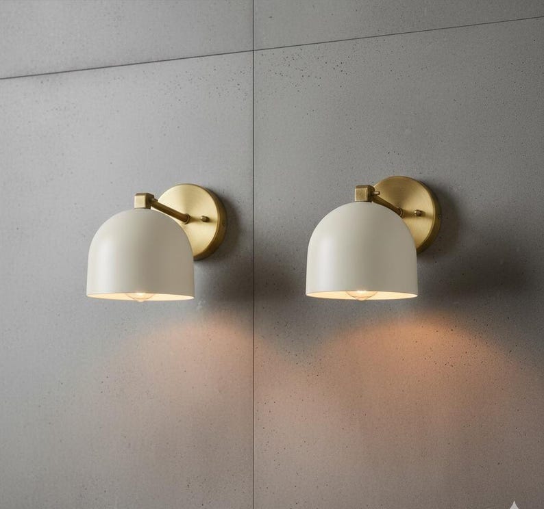 May include: Two matching wall sconces with a cream-colored, dome-shaped shade and a brass-colored circular backplate. The sconces are mounted on a gray wall, and the light from the bulbs creates a warm glow.