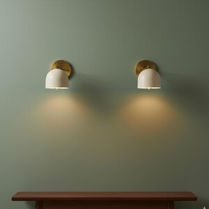 May include: Two wall sconces with a brass finish and off-white, dome-shaped shades. The sconces are mounted on a muted green wall, casting a warm glow downwards. The sconces are a modern lighting fixture.
