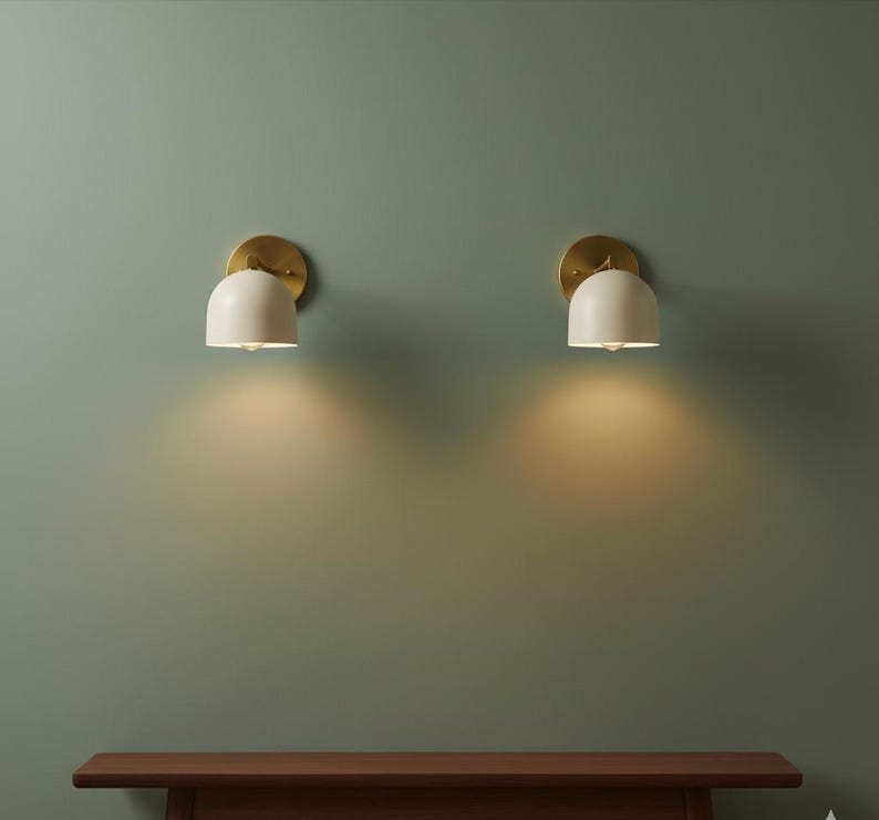 May include: Two wall sconces with a brass finish and off-white, dome-shaped shades. The sconces are mounted on a muted green wall, casting a warm glow downwards. The sconces are a modern lighting fixture.