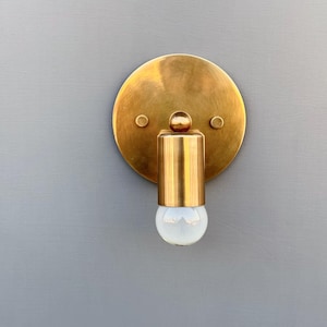 May include: A brass wall sconce with a round backplate and a cylindrical light fixture. The sconce has a warm, golden hue and a frosted glass bulb. The fixture is mounted on a gray wall.