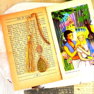 Könnte beinhalten: Ein handgefertigtes Lesezeichen mit einer goldenen Kette und einem tropfenförmigen Anhänger mit einem grün-goldenen Design. Das Lesezeichen befindet sich in einem offenen Buch mit französischem Text und einer farbenfrohen Illustration.