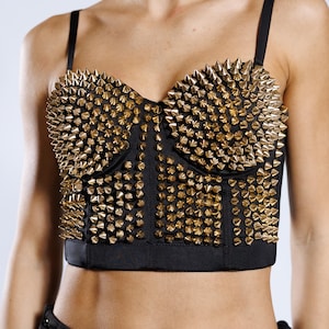 Könnte beinhalten: Schwarzes Bustier-Top mit goldfarbenen Kegelnieten. Das Top hat verstellbare Träger und ein strukturiertes Oberteil. Die Nieten sind dicht gepackt und bedecken die gesamte Vorderseite. Ein auffälliges Kleidungsstück.