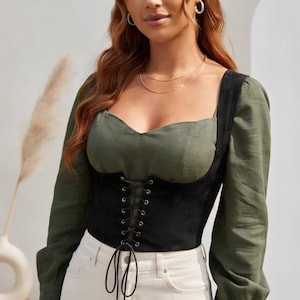 May include: An olive green long-sleeve top featuring a sweetheart neckline and a black corset-style bodice overlay. The black section includes a front lace-up detail with gold-toned metal eyelets and long black cord ties hanging at the waist.