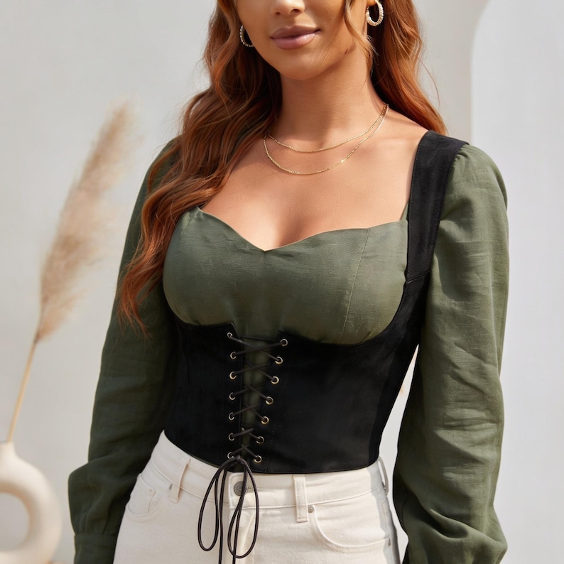 May include: An olive green long-sleeve top featuring a sweetheart neckline and a black corset-style bodice overlay. The black section includes a front lace-up detail with gold-toned metal eyelets and long black cord ties hanging at the waist.