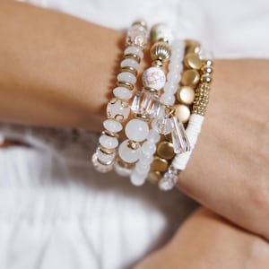 May include: A collection of white and gold beaded bracelets. The bracelets feature various designs, including round beads, gold accents, and clear crystal charms. Some bracelets have floral patterns. The bracelets are stacked together.