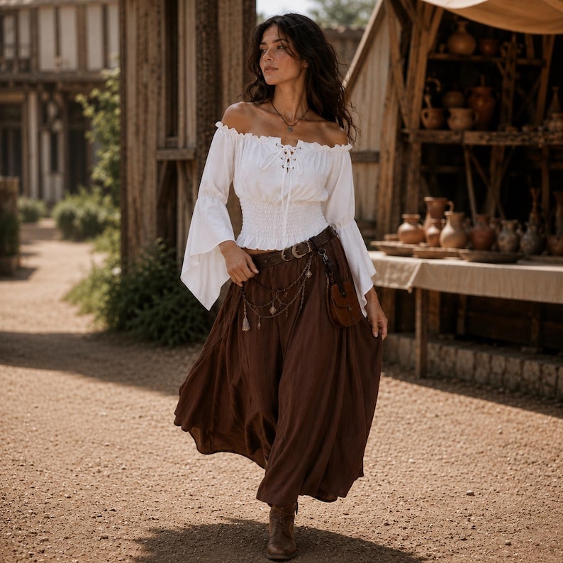 K&ouml;nnte beinhalten: Eine wei&szlig;e schulterfreie Bluse mit langen, ausgestellten &Auml;rmeln, Schn&uuml;rung vorne und gesmokter Taille, kombiniert mit einem langen, flie&szlig;enden dunkelbraunen Rock. Das Outfit umfasst einen braunen Lederg&uuml;rtel mit kleiner Tasche und Kette.