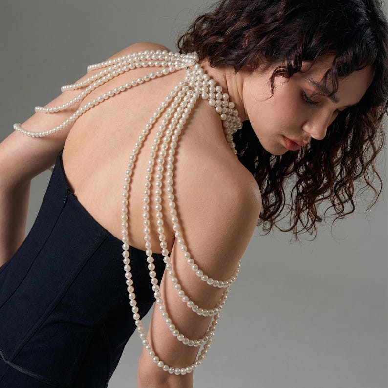 Pode incluir: Uma mulher vestindo um vestido preto com joias de p&eacute;rolas nos ombros e bra&ccedil;os. As joias consistem em v&aacute;rios fios de pequenas p&eacute;rolas redondas, esbranqui&ccedil;adas, caindo sobre os ombros e bra&ccedil;os. As p&eacute;rolas criam um efeito elegante e em camadas.