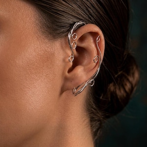 May include: A silver-toned ear cuff featuring a delicate wire-wrapped design that follows the outer curve of the ear. The piece consists of thin, polished metal strands that form multiple small, circular loops and sweeping lines along the cartilage.