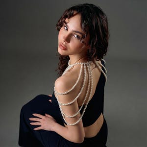 Pode incluir: Uma mulher vestindo um vestido preto com um colar de p&eacute;rolas nos ombros. O colar &eacute; feito de v&aacute;rios fios de p&eacute;rolas que caem sobre os ombros e as costas. O fundo &eacute; cinza liso. A mulher olha por cima do ombro.