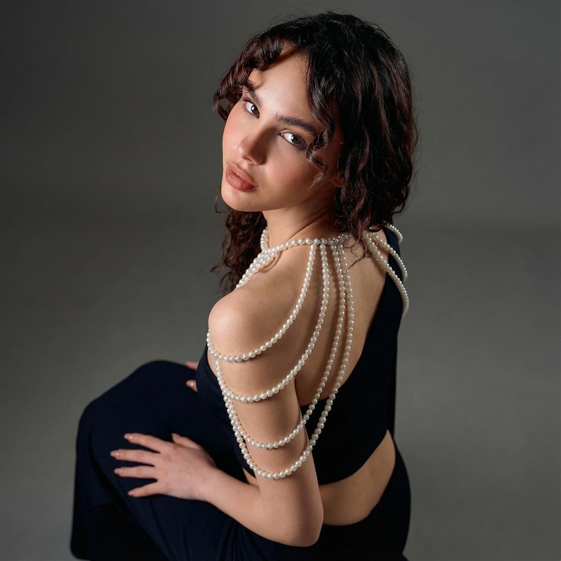 Pode incluir: Uma mulher vestindo um vestido preto com um colar de p&eacute;rolas nos ombros. O colar &eacute; feito de v&aacute;rios fios de p&eacute;rolas que caem sobre os ombros e as costas. O fundo &eacute; cinza liso. A mulher olha por cima do ombro.
