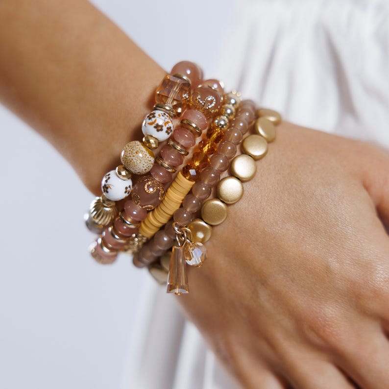 May include: A stack of beaded bracelets in shades of gold, brown, and cream. The bracelets feature various bead styles, including round, disc, and patterned beads. A charm with a crystal pendant hangs from one bracelet.