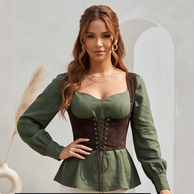May include: A long-sleeved olive green blouse featuring a sweetheart neckline and a dark brown suede-like corset-style bodice overlay. The bodice includes a central lace-up detail with metal eyelets and long brown cord ties hanging at the waist.