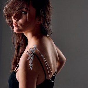 May include: A silver shoulder piece featuring a floral and leaf design, embellished with clear crystals. The shoulder jewellery hangs from a delicate chain. The piece is worn over a black top.