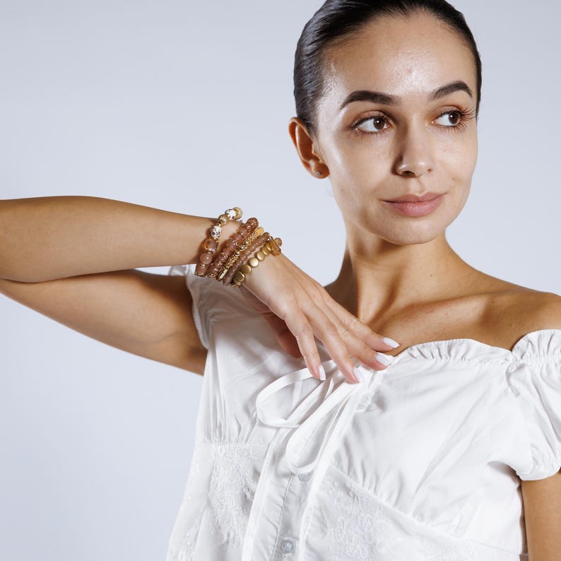 May include: A collection of gold and brown beaded bracelets. The bracelets are stacked on a wrist. The bracelets have different textures and shapes, including round beads and flat, circular pieces. The background is a solid, light color.