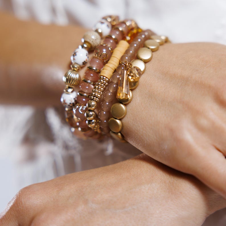 May include: A collection of beaded bracelets in shades of gold, pink, and cream. The bracelets feature various textures, including round beads, disc-shaped beads, and a wooden accent. A small, dangling charm adds a touch of elegance.