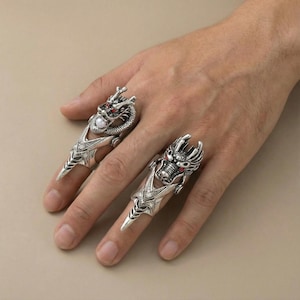 May include: Two silver-colored dragon rings with red and white accents. The rings feature detailed dragon heads and extend down the finger. One ring has a pearl accent. The rings are on a hand.