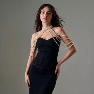Pode incluir: Um vestido preto sem al&ccedil;as com um colar de p&eacute;rolas drapeado sobre os ombros. O colar &eacute; feito de v&aacute;rios fios de pequenas p&eacute;rolas redondas, brancas. O vestido tem um corpete justo e uma saia longa.
