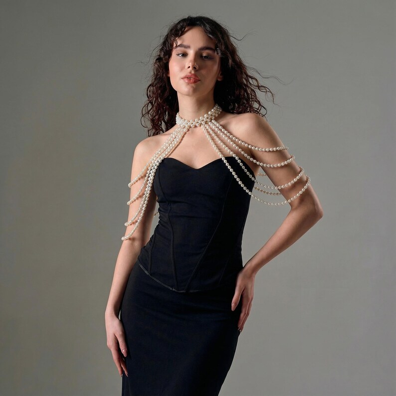 Pode incluir: Um vestido preto sem al&ccedil;as com um colar de p&eacute;rolas drapeado sobre os ombros. O colar &eacute; feito de v&aacute;rios fios de pequenas p&eacute;rolas redondas, brancas. O vestido tem um corpete justo e uma saia longa.