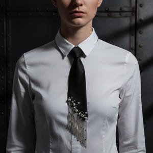 Puede incluir: Una corbata negra adornada con perlas y cadenas plateadas colgantes. La corbata se lleva sobre una camisa blanca. El fondo es una superficie oscura y texturizada.