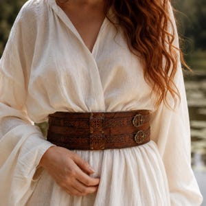 May include: A wide brown leather corset belt featuring intricate embossed Celtic knot patterns, two horizontal straps, and two ornate brass-toned metal buckles. The belt is worn over a long-sleeved, off-white, textured linen-style blouse.