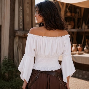 K&ouml;nnte beinhalten: Eine wei&szlig;e schulterfreie Bluse mit langen, ausgestellten Glocken&auml;rmeln und einer gerafften, elastischen gesmokten Taille. Das Kleidungsst&uuml;ck hat einen ger&uuml;schten Ausschnitt und einen ger&uuml;schten Saum, was f&uuml;r eine taillierte Passform sorgt.