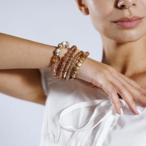 May include: A collection of beaded bracelets in shades of gold, brown, and pink. The bracelets feature various textures, including round beads, faceted beads, and gold-colored metal accents. The bracelets are stacked on a wrist.