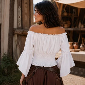 K&ouml;nnte beinhalten: Eine wei&szlig;e schulterfreie Bluse mit langen, ausgestellten Glocken&auml;rmeln und einer gerafften, elastischen gesmokten Taille. Das Kleidungsst&uuml;ck hat einen ger&uuml;schten Ausschnitt und eine kurze Silhouette, kombiniert mit einem dunkelbraunen Rock mit Lederg&uuml;rtel.