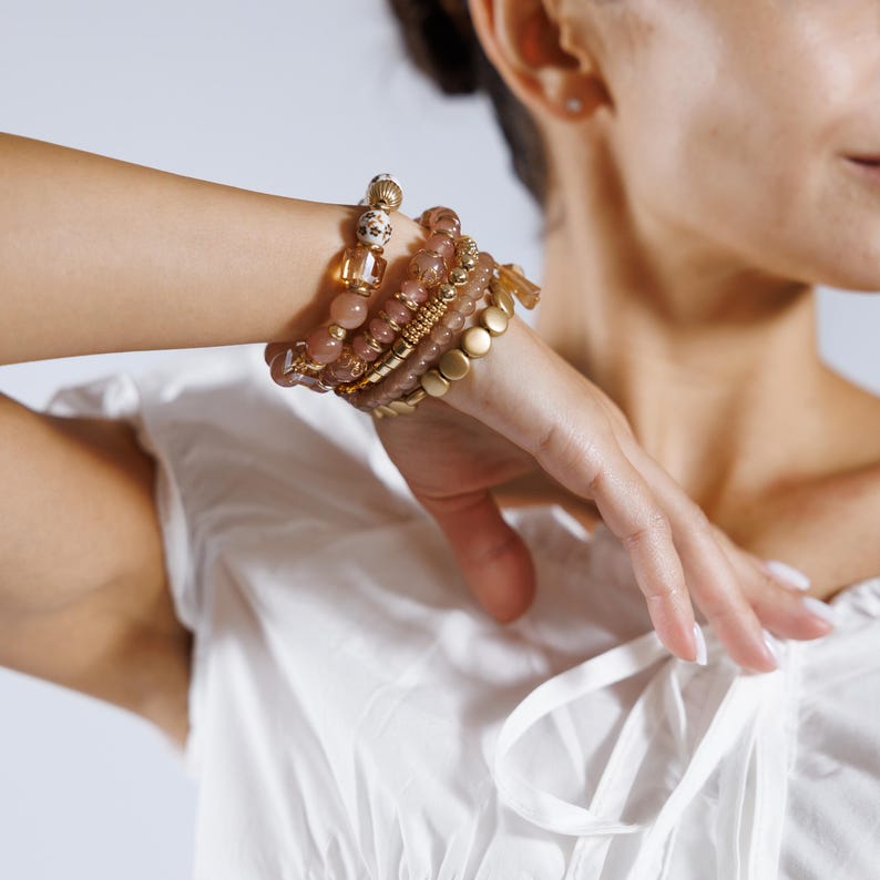 May include: A collection of beaded bracelets in shades of gold, pink, and brown adorns a wrist. The bracelets feature various textures, including smooth beads, textured gold accents, and a leopard print bead. The bracelets are stacked together for a layered look.