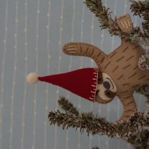 May include: A brown felt sloth wearing a red Santa hat with a white pom-pom, hanging from a green artificial Christmas tree.