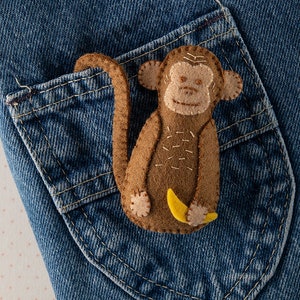 May include: A brown felt monkey with a yellow felt banana in its hand, sitting in a blue denim pocket.