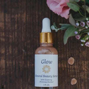 May include: A glass bottle of Glow Mineral Beauty Serum with a white label and gold accents. The label includes the text "Glow" and "With Evening Primrose Extract". The bottle is 1 fl oz. Pink flowers and greenery are in the background.