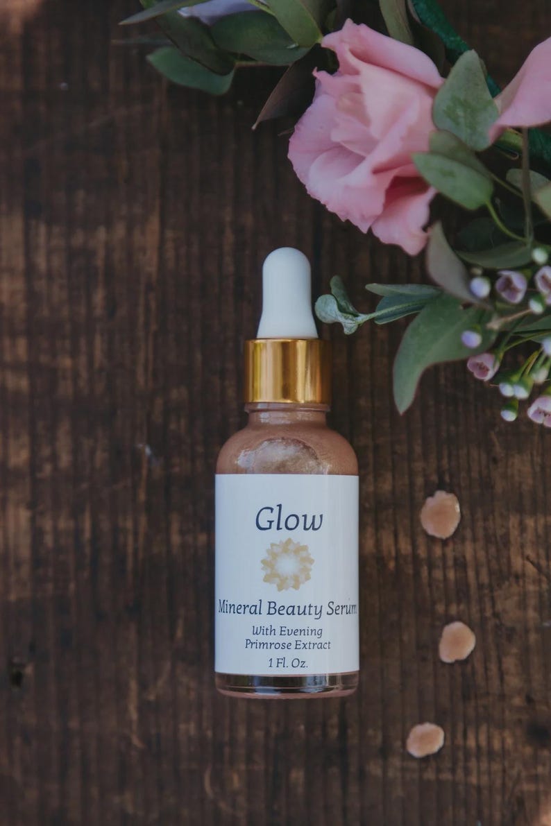May include: A glass bottle of Glow Mineral Beauty Serum with a white label and gold accents. The label includes the text "Glow" and "With Evening Primrose Extract". The bottle is 1 fl oz. Pink flowers and greenery are in the background.