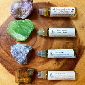 May include: An overhead shot features four facial serum roll-on bottles with crystal rollers, each labeled with a different name. The bottles are accompanied by raw gemstones in shades of purple, green, blue, and brown, all arranged on a wooden surface.