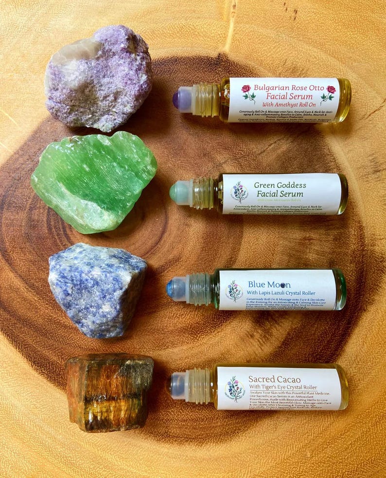 May include: An overhead shot features four facial serum roll-on bottles with crystal rollers, each labeled with a different name. The bottles are accompanied by raw gemstones in shades of purple, green, blue, and brown, all arranged on a wooden surface.
