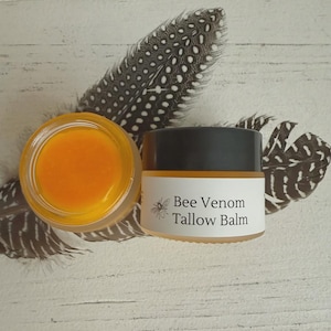Op de afbeelding: Twee kleine potjes balsem, waarvan één open, met een oranje-gele substantie. Het andere potje is gesloten met een zwart deksel en een etiket met de tekst "Bee Venom Tallow Balm" met een bijenillustratie. De potjes staan op een wit houten oppervlak met veren.