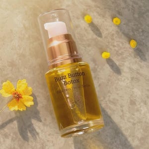 May include: A clear glass pump bottle containing golden facial serum with the text Buzz Button Botox Paracress Facial Serum 1 FL OZ printed on the front. The bottle has a metallic gold collar and a clear cap, surrounded by small yellow flowers.