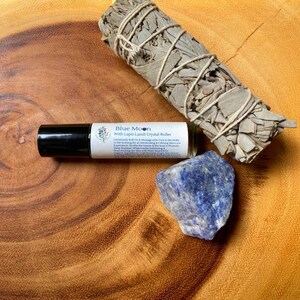 May include: A collection of spiritual items on a wooden surface. The items include a roll-on bottle labeled "Blue Moon With Lapis Lazuli Crystal Roller", a bundle of dried sage, and a piece of blue lapis lazuli crystal.