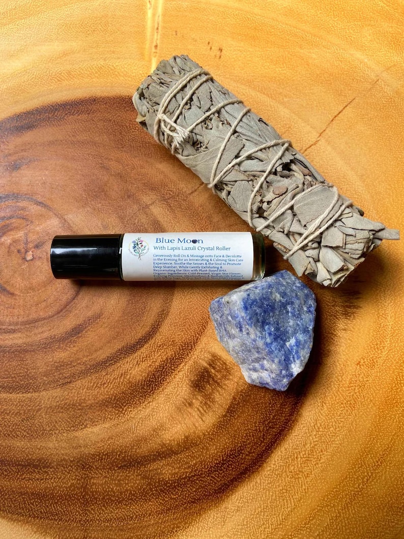 May include: A collection of spiritual items on a wooden surface. The items include a roll-on bottle labeled "Blue Moon With Lapis Lazuli Crystal Roller", a bundle of dried sage, and a piece of blue lapis lazuli crystal.