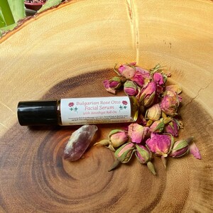 May include: A roll-on bottle of Bulgarian Rose Otto Facial Serum with Amethyst Roll On, a purple crystal, and dried pink rose buds are arranged on a wooden surface. The serum bottle has a black cap and a white label with text and floral graphics.