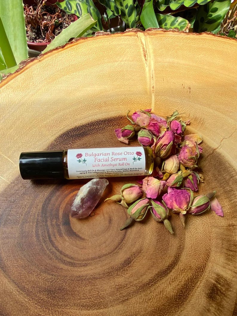 May include: A roll-on bottle of Bulgarian Rose Otto Facial Serum with Amethyst Roll On, a purple crystal, and dried pink rose buds are arranged on a wooden surface. The serum bottle has a black cap and a white label with text and floral graphics.
