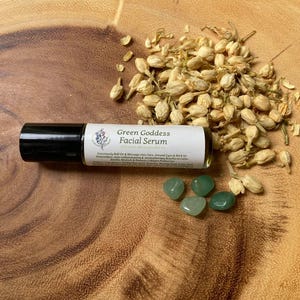 May include: A roll-on facial serum labeled "Green Goddess Facial Serum" with a black cap, alongside green gemstones and dried flower buds. The serum's label includes text about its benefits. The items are arranged on a wooden surface.