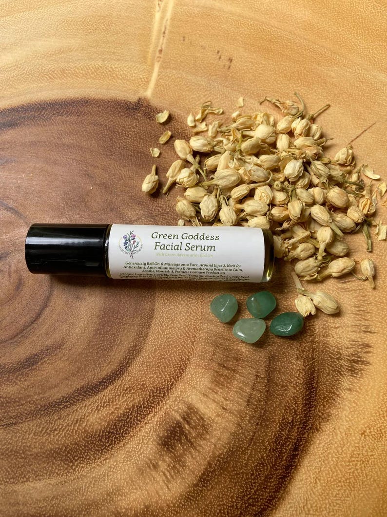 May include: A roll-on facial serum labeled "Green Goddess Facial Serum" with a black cap, alongside green gemstones and dried flower buds. The serum's label includes text about its benefits. The items are arranged on a wooden surface.