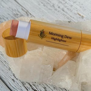 Puede incluir: Un lápiz de resaltador de bambú con el texto "Morning Dew Highlighter" impreso en el lateral. El resaltador es de color rosa claro y está parcialmente extendido de su tubo de bambú. La tapa también es de bambú.
